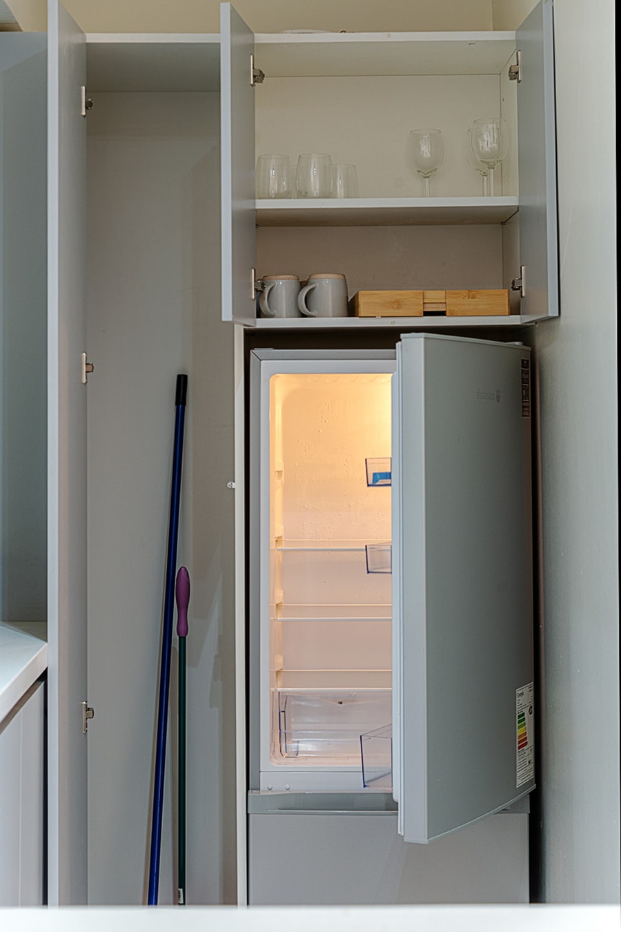 Refrigerador abierto dentro del mueble de la cocina, con repisa alta de copas y tazas y utensilios de aseo al costado