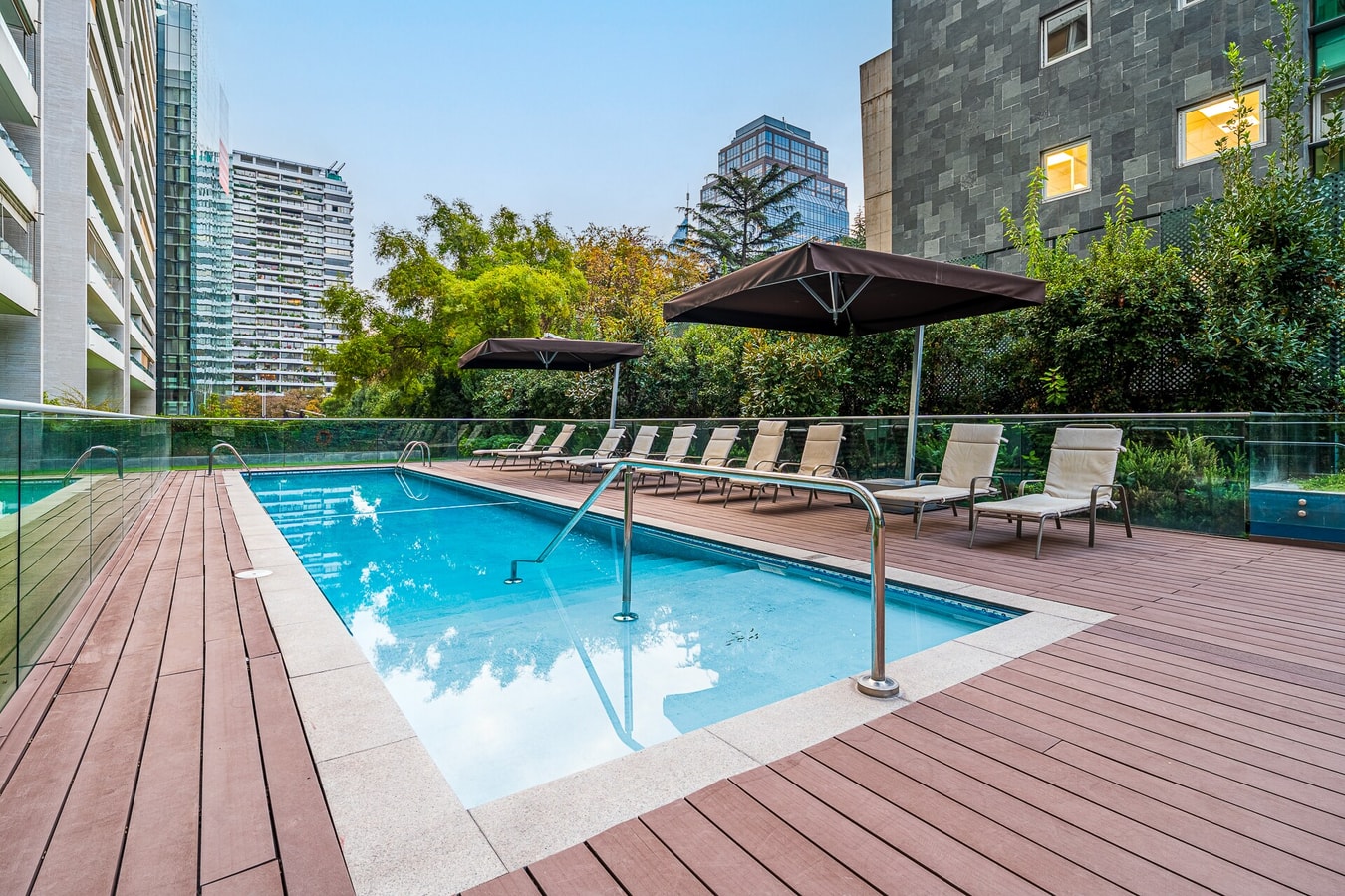 Piscina del edificio con deck de madera y camastros, rodeada de vegetación.