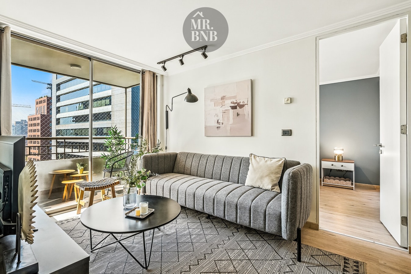 Living con sofá gris, mesa de centro y ventanales amplios con vista hacia los edificios.