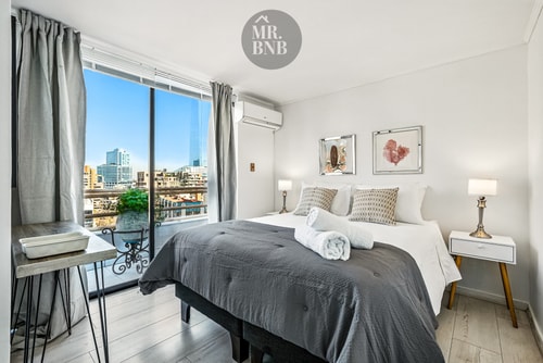 Dormitorio con cama, veladores, escritorio y ventanal con aire acondicionado