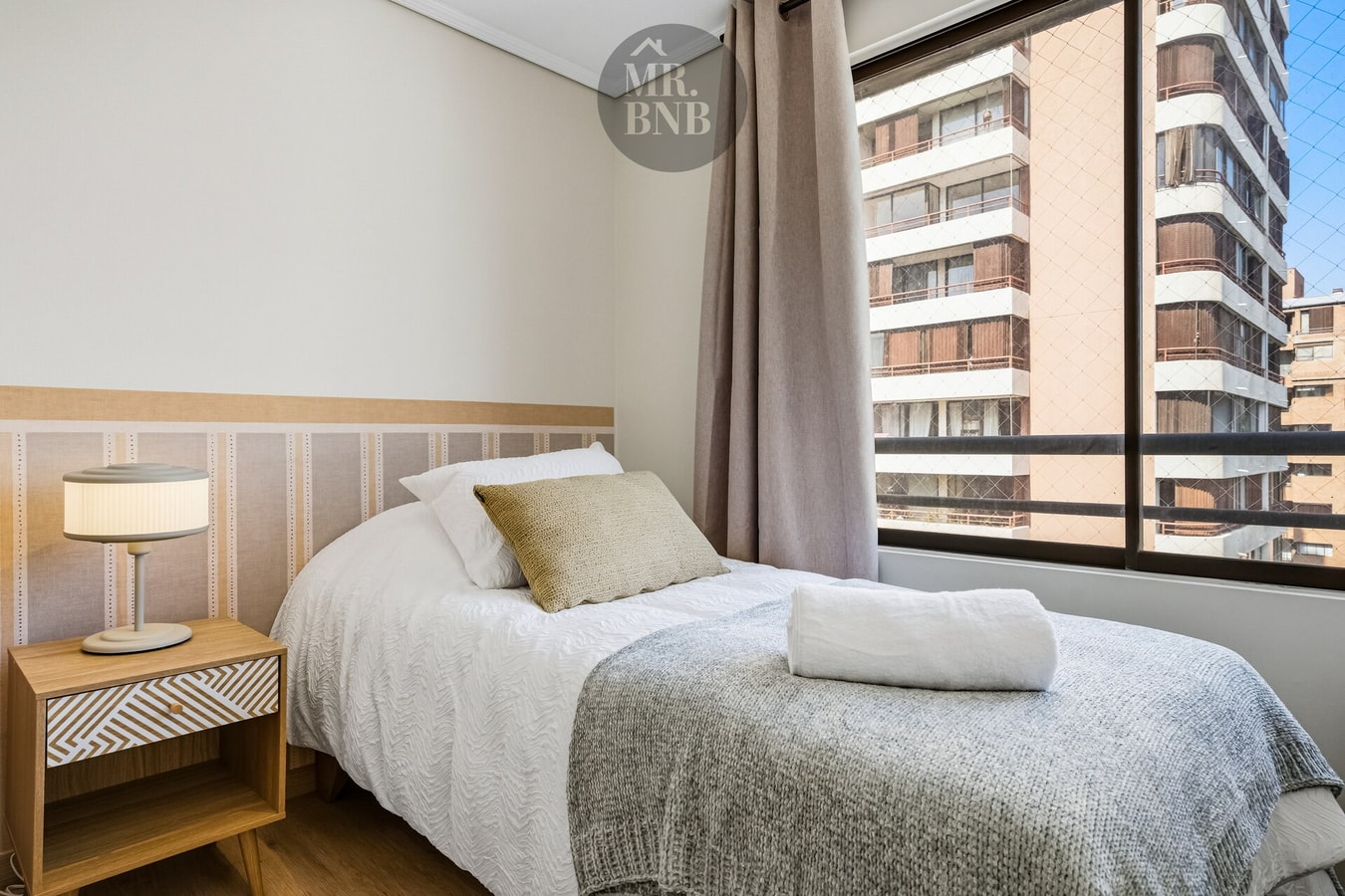 Dormitorio individual con cama, velador y ventana con vista a edificios