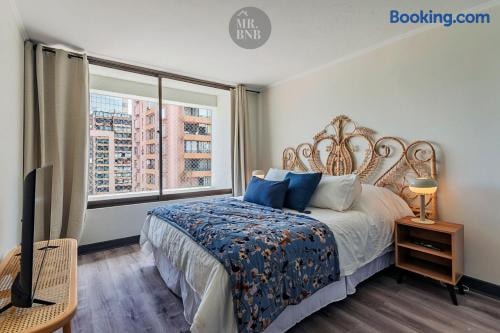 Dormitorio con cama, cabecero de mimbre y ventanal con vista a edificios.