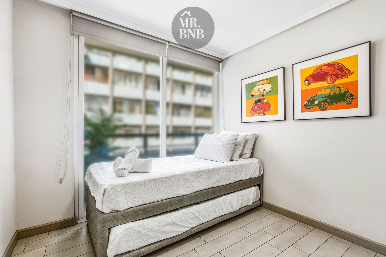 Dormitorio con dos camas, cuadros de autos y ventanal con vista a edificios.