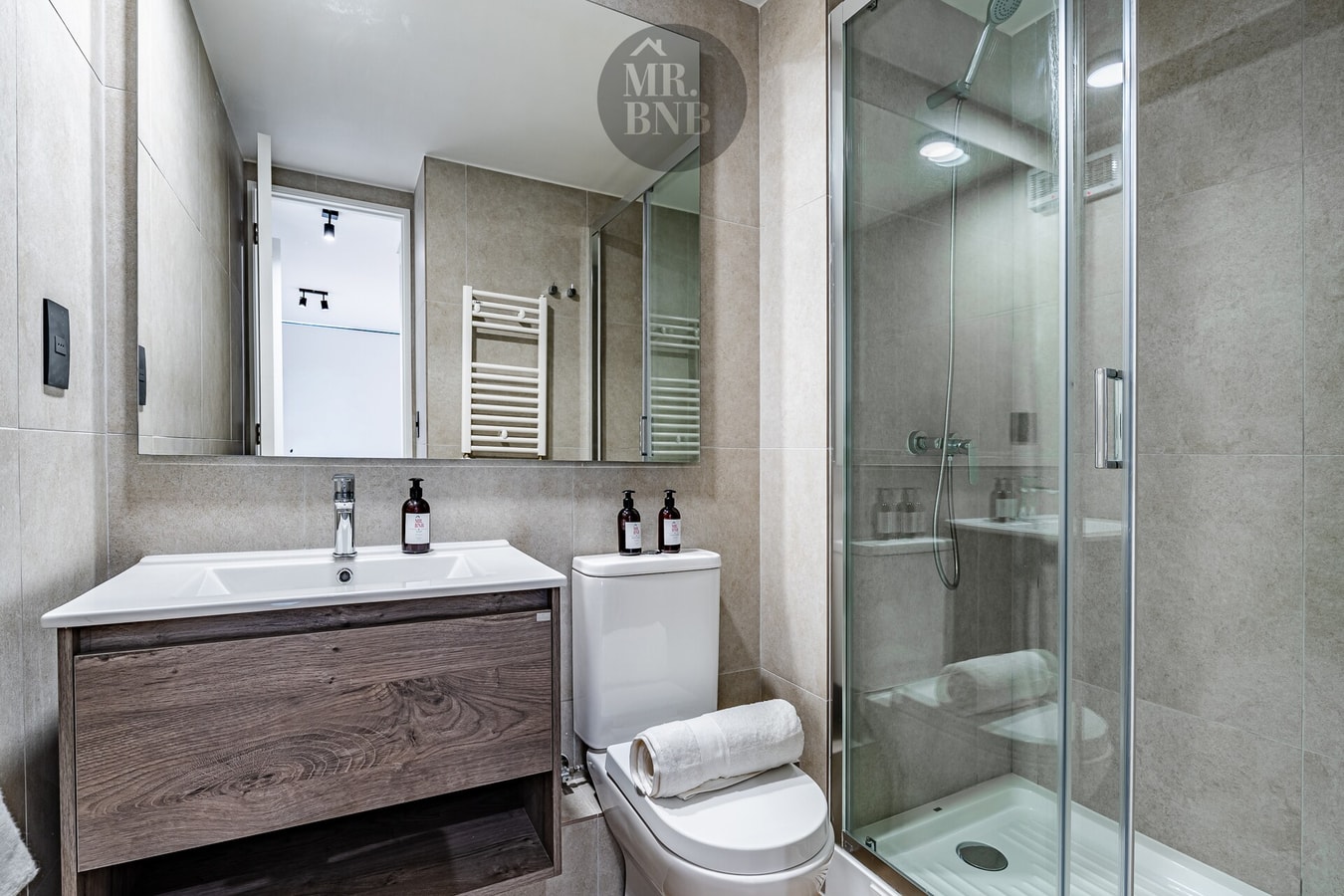 Baño con ducha de vidrio, mueble vanitorio de madera oscura y toallero.