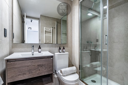 Baño con ducha de vidrio, mueble vanitorio de madera oscura y toallero.