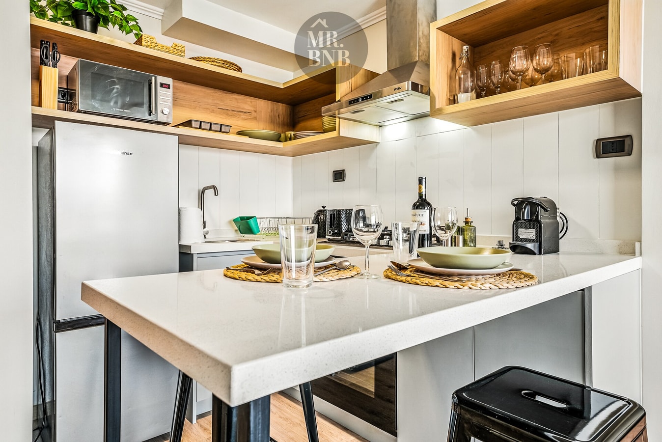 Cocina con isla barra, microondas, vajilla y cafetera sobre muebles de madera.