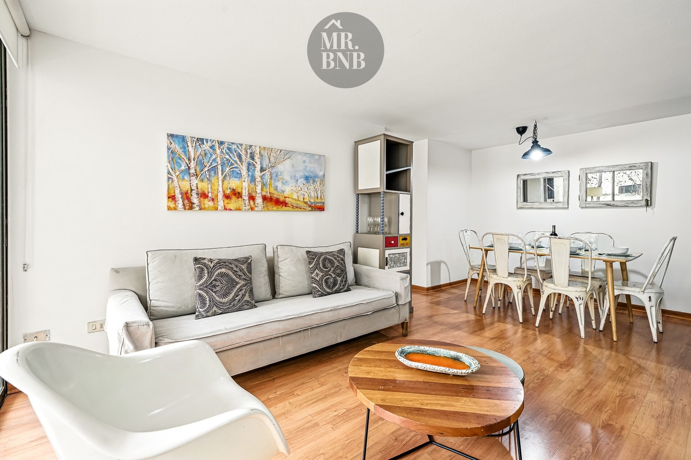 Living con sofá gris, sillón blanco y comedor con sillas metálicas al fondo