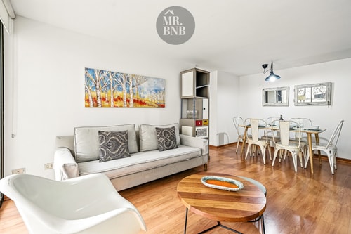 Living con sofá gris, sillón blanco y comedor con sillas metálicas al fondo