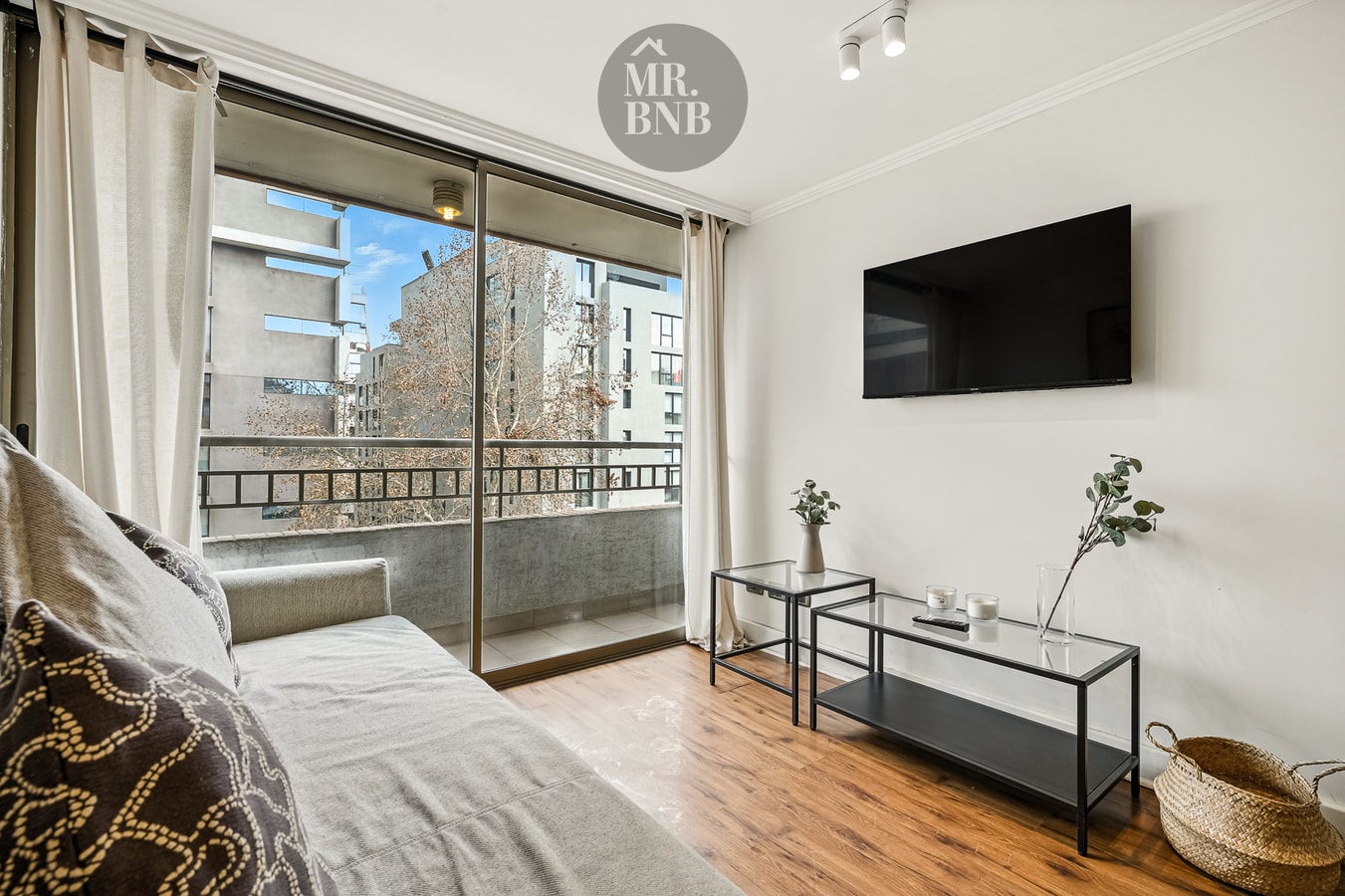 Dormitorio con cama y velador, ventanal con cortinas, televisor en la pared y mesa auxiliar