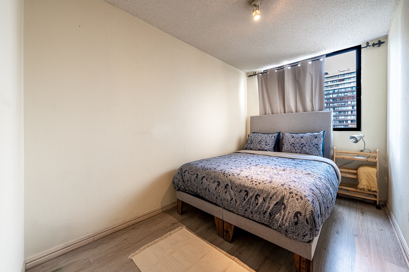 Dormitorio con cama, mesa de noche y ventana con cortina y vista al edificio vecino.