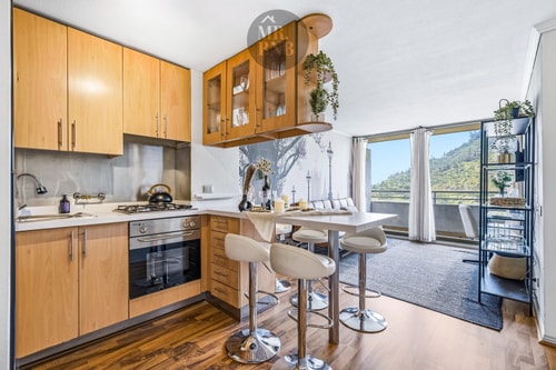 Cocina de madera en L con barra alta y sillas, vista hacia el living con ventanales.