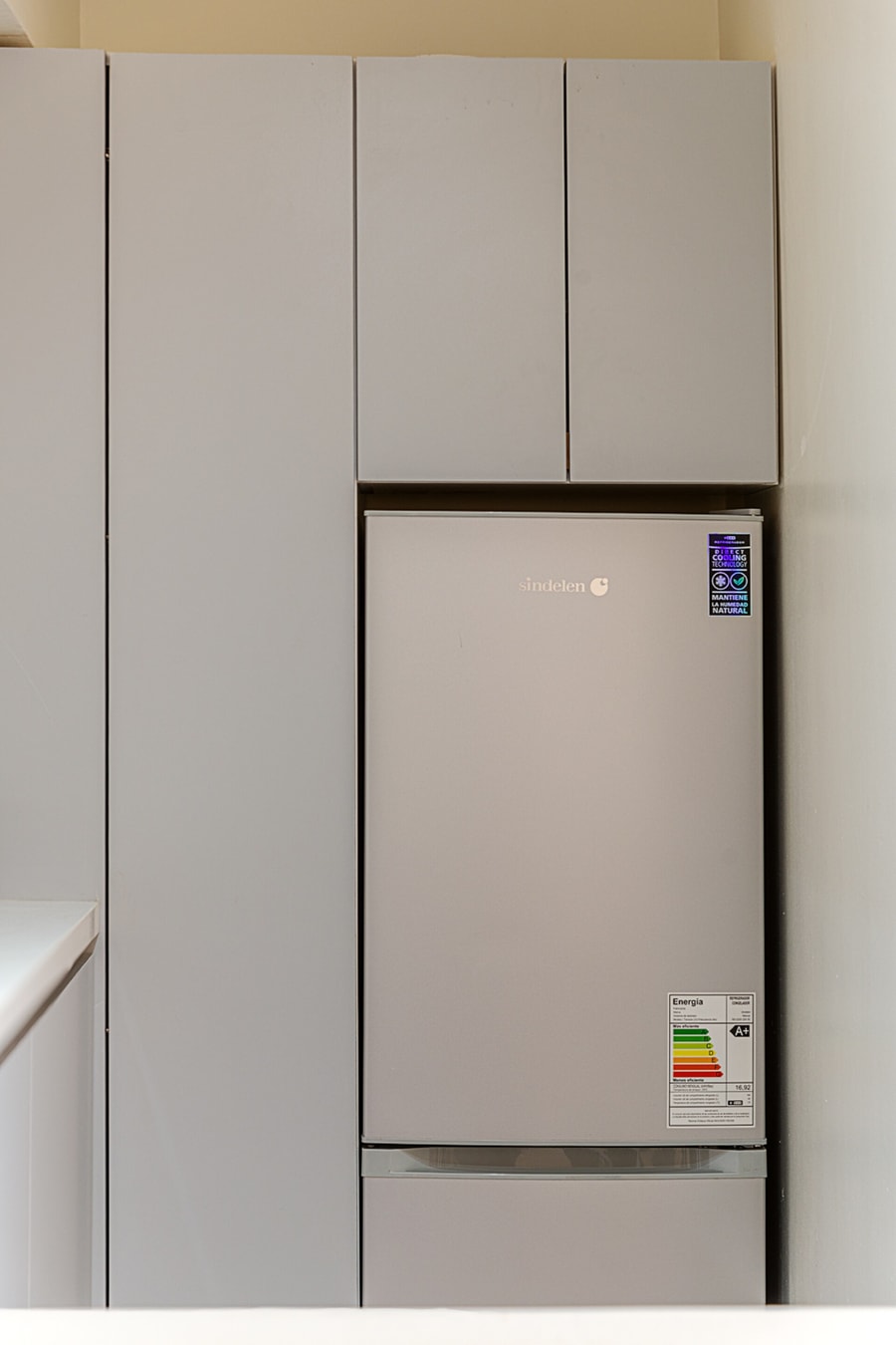 Refrigerador empotrado entre muebles de cocina grises, con puertas altas de despensa sobre él