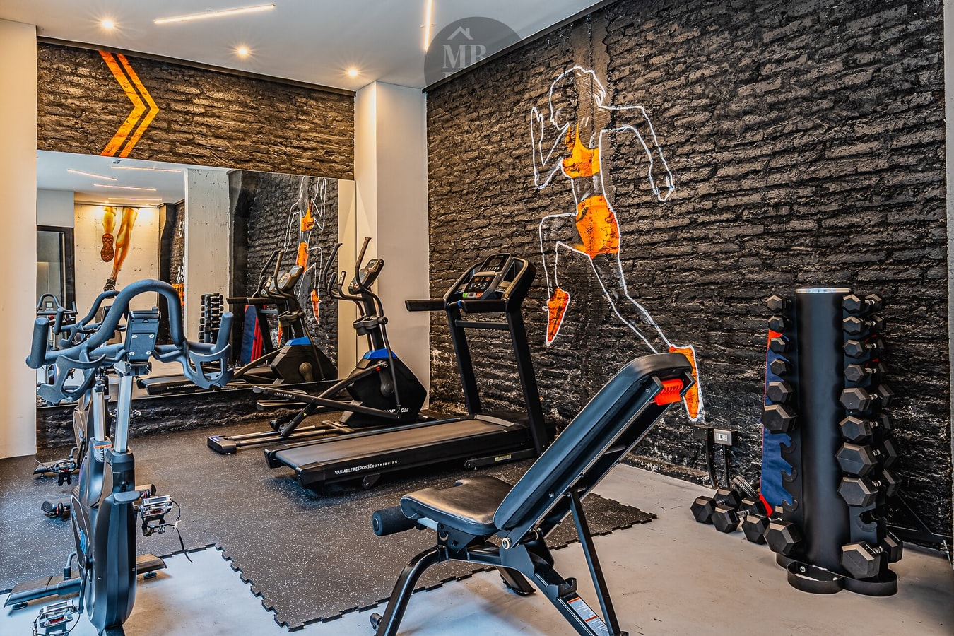 Gimnasio del edificio con bicicletas estáticas, elíptica, trotadora, banco de pesas y mancuernas, sobre muro de piedra