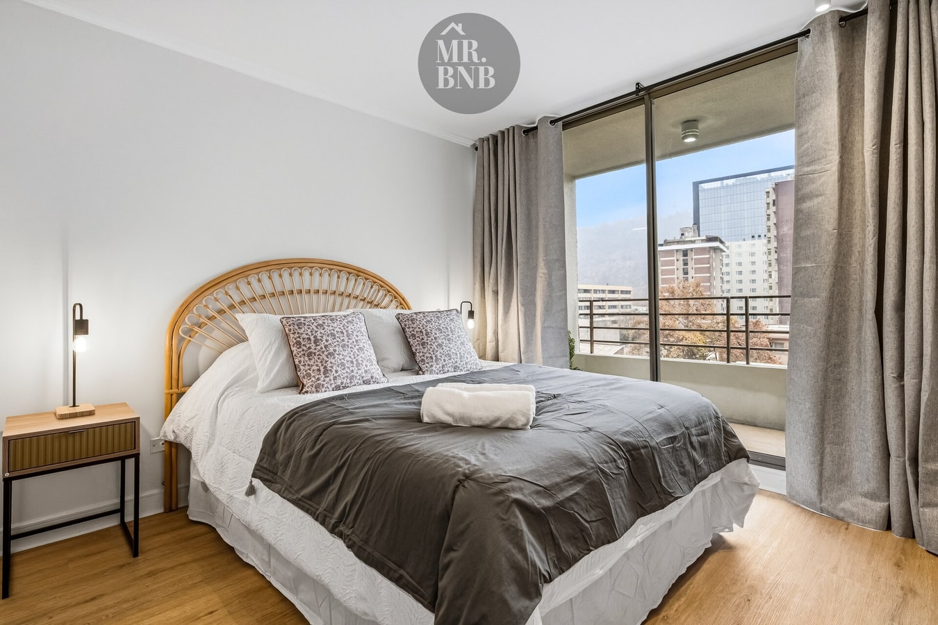 Dormitorio con cama matrimonial, cabecero de mimbre y ventanal con vista a edificios