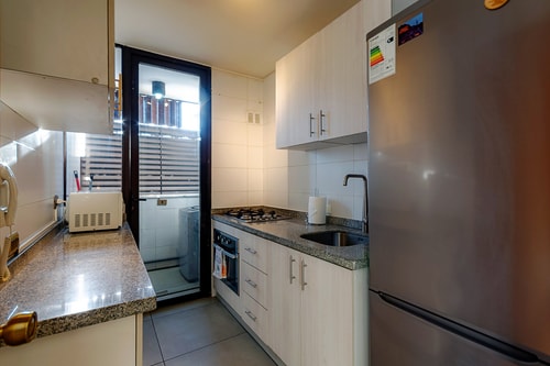 Cocina con refrigerador, lavaplatos y muebles blancos junto a ventana lateral.
