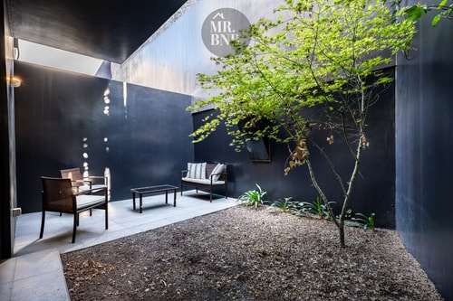 Patio interior privado con muros oscuros, árbol, sillones, mesa de centro y deck de piedra
