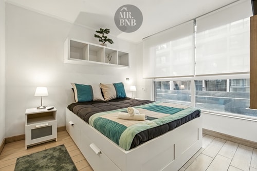 Dormitorio con cama, velador con lámpara y cortina de enrollar en la ventana.