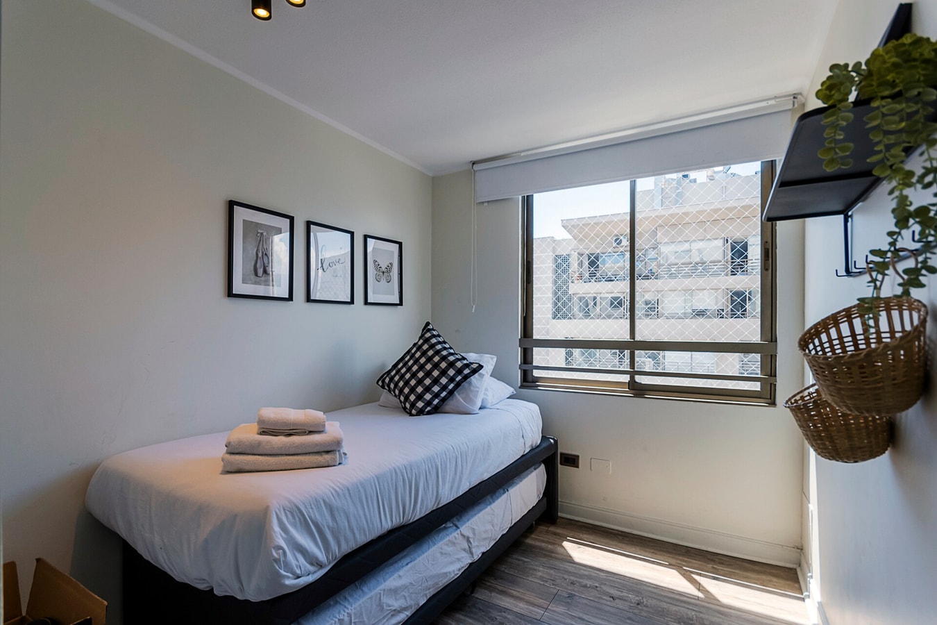 Dormitorio con cama, cuadros en la pared, ventana con vista a edificios y repisa con plantas