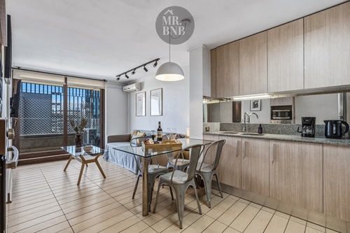 Living comedor integrado a la cocina, con mesa de vidrio, sillas metálicas y aire acondicionado en el techo.