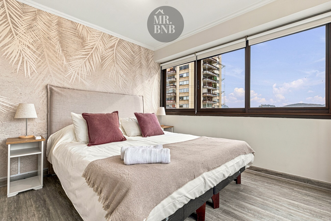 Dormitorio con cama de dos plazas, papel mural con estampado de hojas, veladores y ventanal.