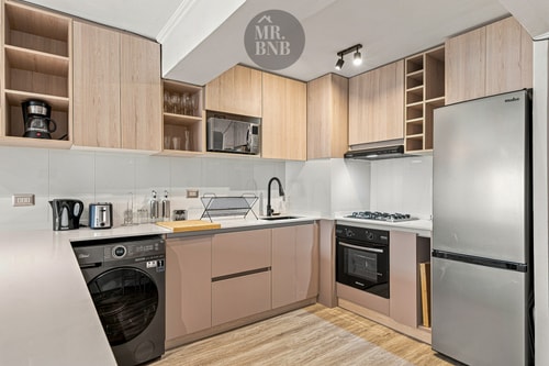 Cocina equipada con muebles en tonos rosa y madera, campana extractora, lavadora bajo encimera y refrigerador.