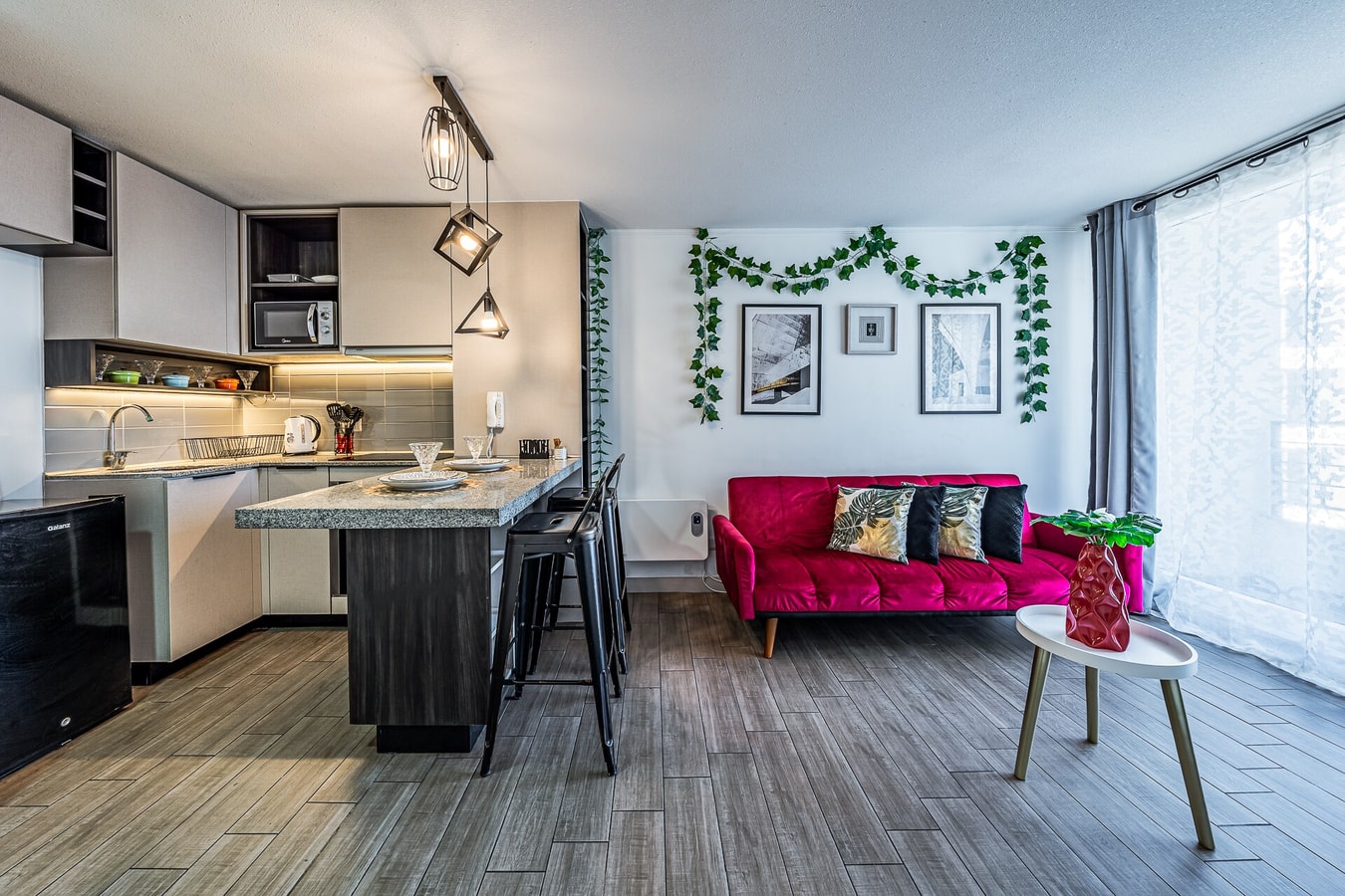 Cocina y living integrados desde otro ángulo, mesón central, sofá rojo y ventana con cortina blanca