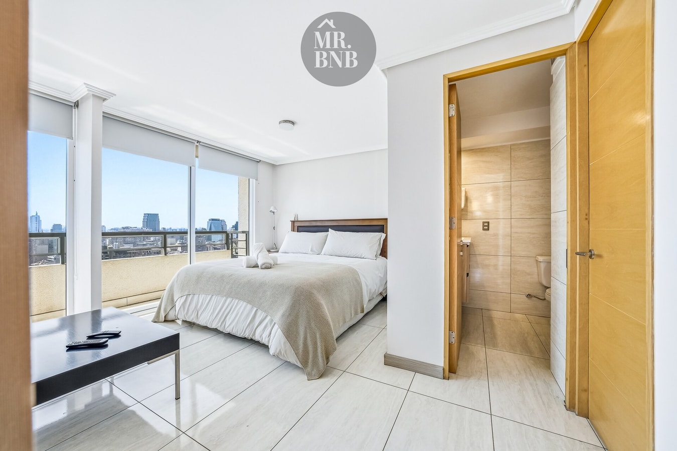 Dormitorio con cama de dos plazas, ventanales con vista a la ciudad y puerta al baño