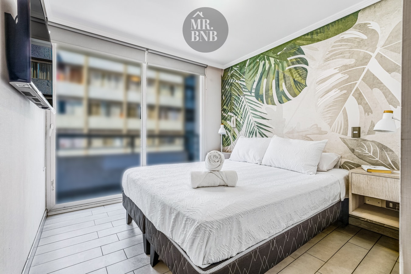 Dormitorio con cama, velador y ventanal con vista a edificios.