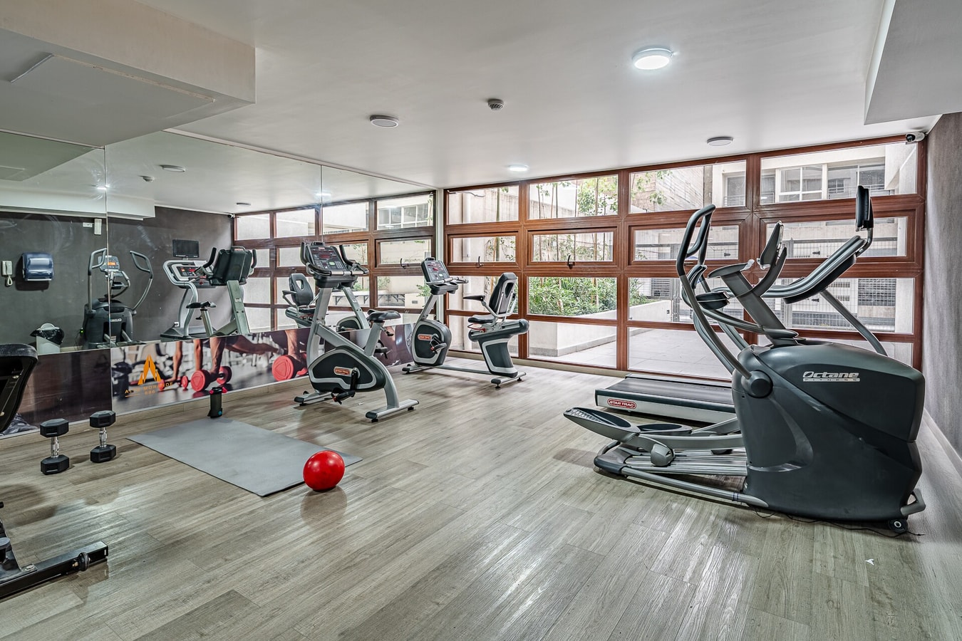 Gimnasio del edificio con máquinas elípticas, pesas y colchoneta frente a ventanales.