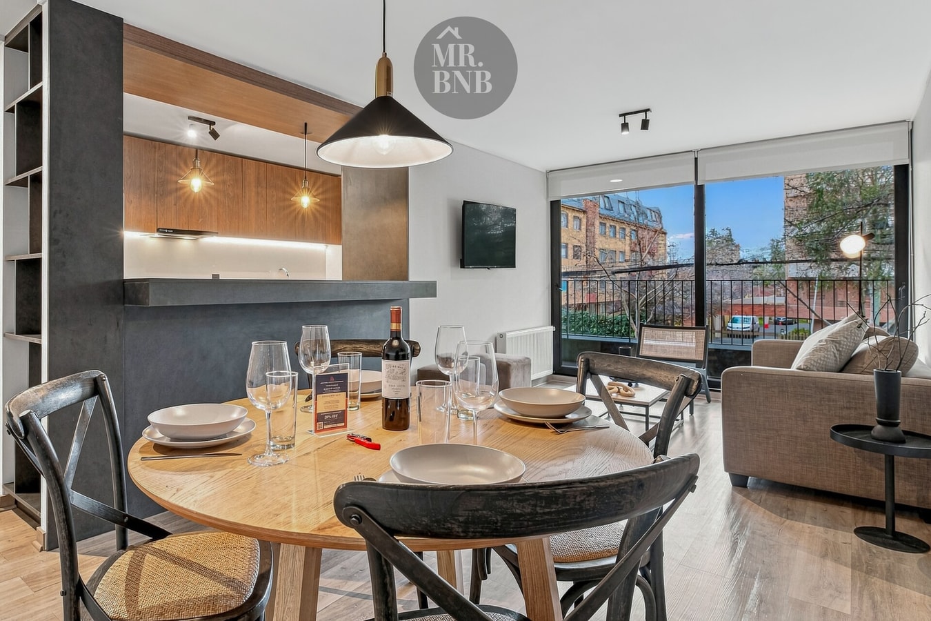 Living comedor con barra de cocina en madera y metal, mesa puesta y sofá gris al fondo