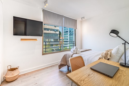 Dormitorio con cama de dos plazas, TV montada, escritorio con notebook y ventana