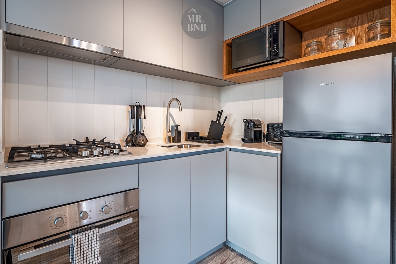 Cocina compacta en L con campana extractora, horno, refrigerador y muebles blancos con cubierta de madera