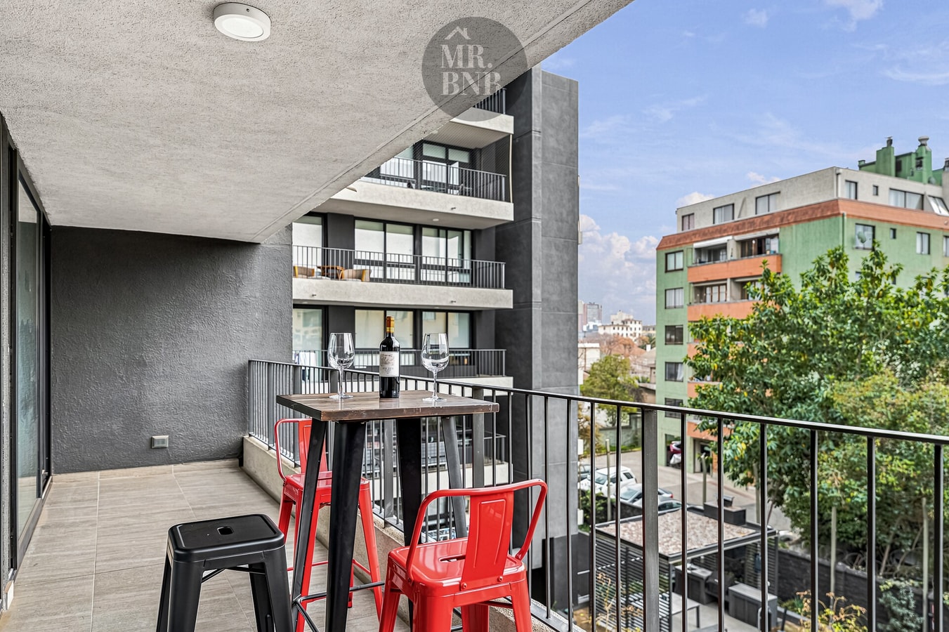Terraza privada con mesa alta y sillas rojas, vista a edificios y arboleda