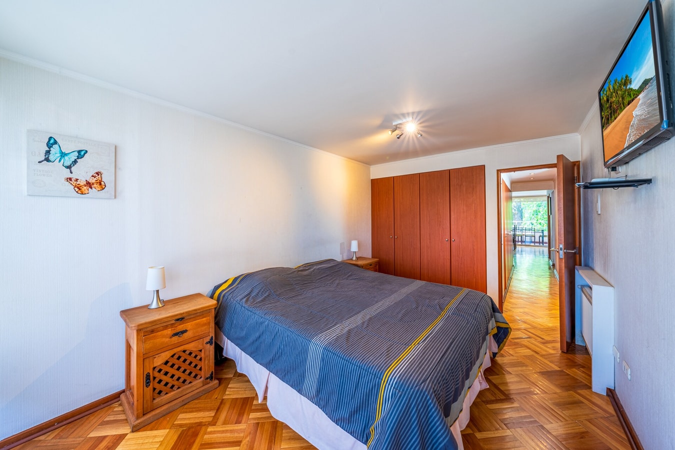 Dormitorio con cama, clóset de madera y televisor, con puerta hacia el pasillo iluminado.