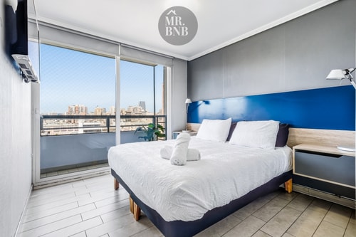 Dormitorio principal con cama, cabecero azul, velador y ventanal con vista a la ciudad