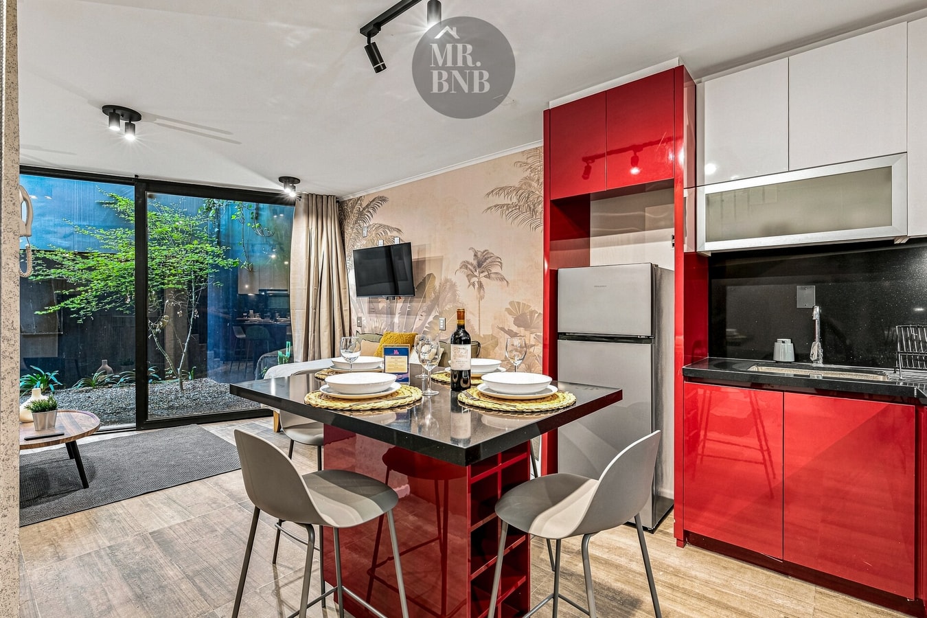 Cocina roja con isla, mesa puesta con vajilla, sillas altas y ventanal hacia el patio