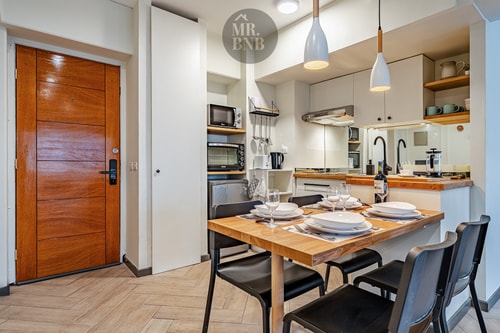 Comedor con mesa de madera y sillas negras, puerta de entrada y cocina al fondo
