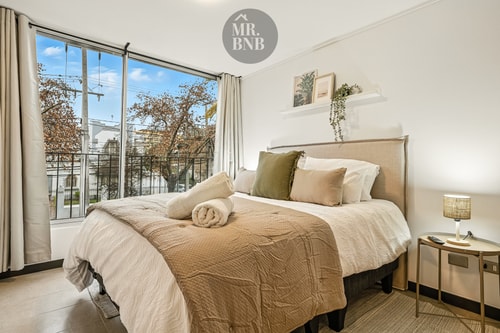 Dormitorio con cama de dos plazas, ropa de cama en tonos beige y ventana con vista a árboles