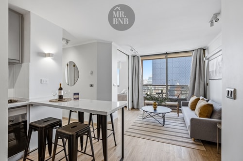 Barra de cocina con banquetas, living con sofá y ventanal con vista a edificios