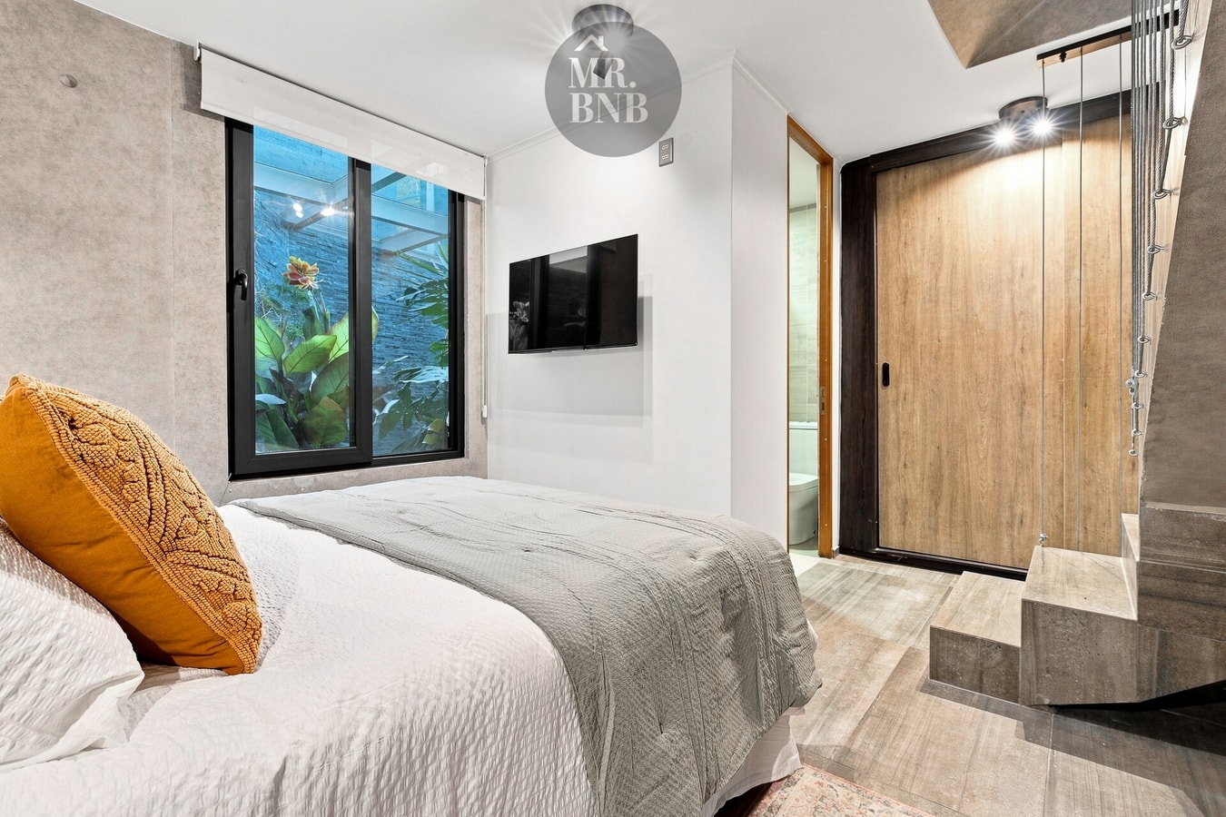 Dormitorio con cama, cojín mostaza, TV y escalera al fondo