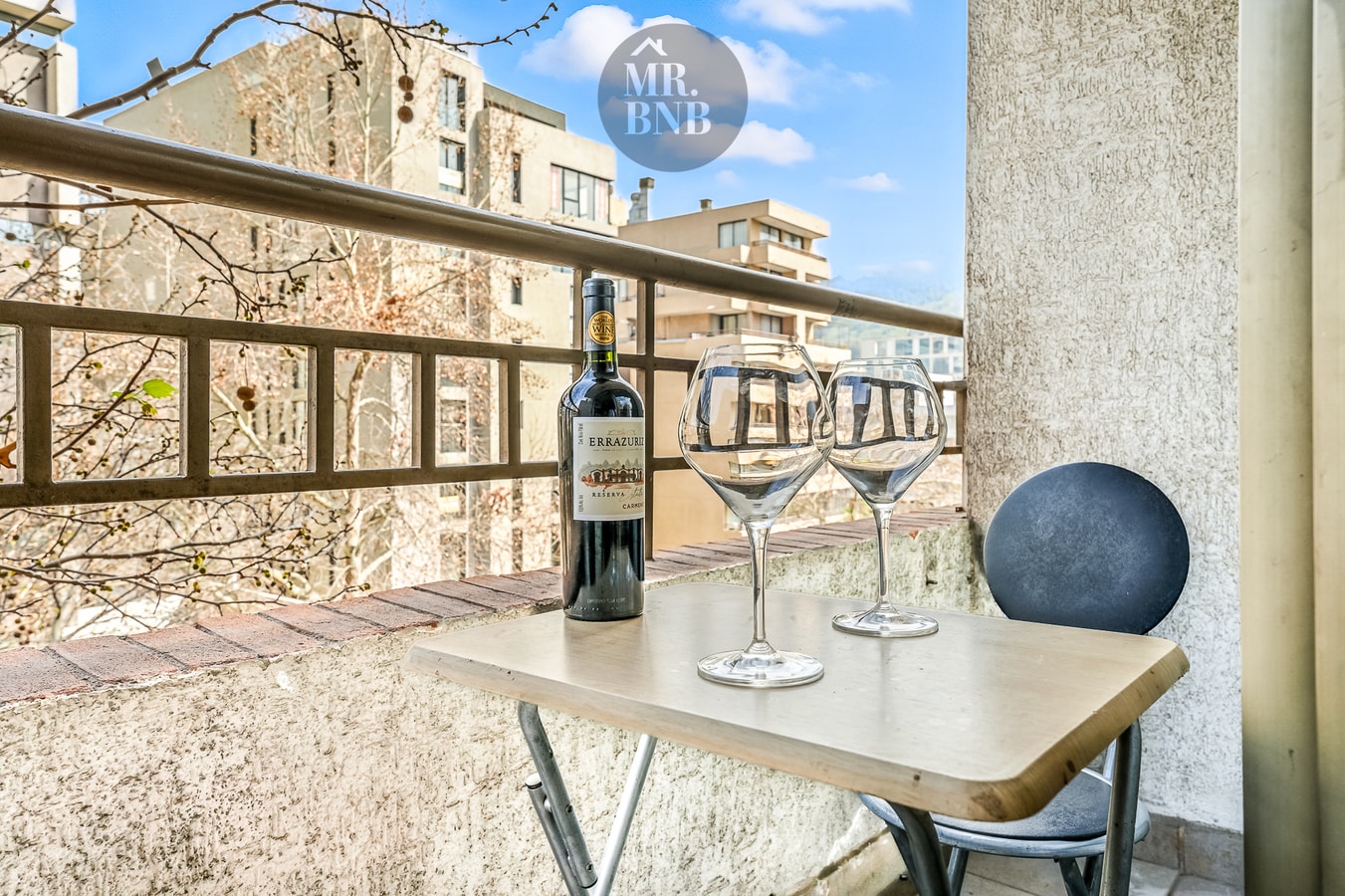 Balcón con mesa, sillas, copas de vino y botella, vista a edificios y árboles