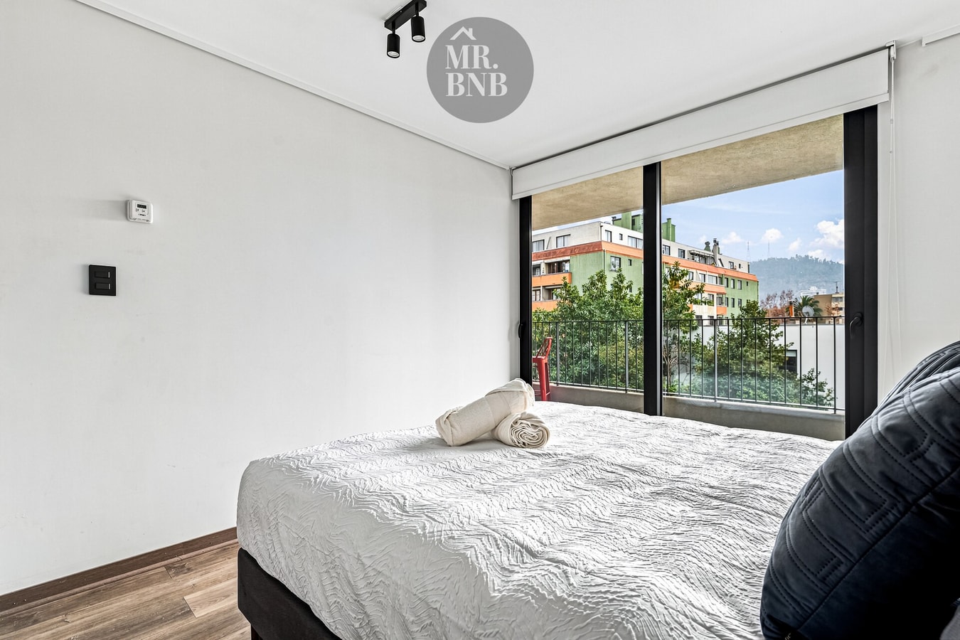 Dormitorio con cama de una plaza junto a ventanal con vista a edificios y cerro