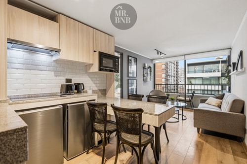 Cocina integrada al living comedor, con muebles de madera clara, sillas de ratán y balcón al fondo