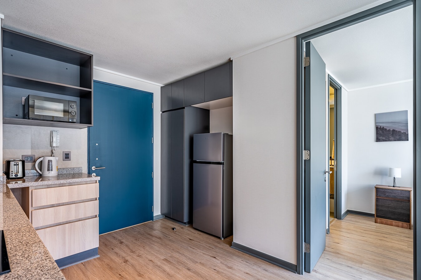 Pasillo de entrada con refrigerador, puerta de acceso azul y vista hacia la cocina y otro ambiente.