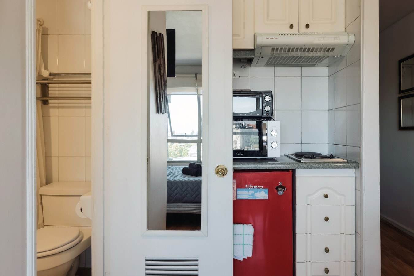 Baño con puerta espejada, kitchenette al fondo con microondas y refrigerador rojo