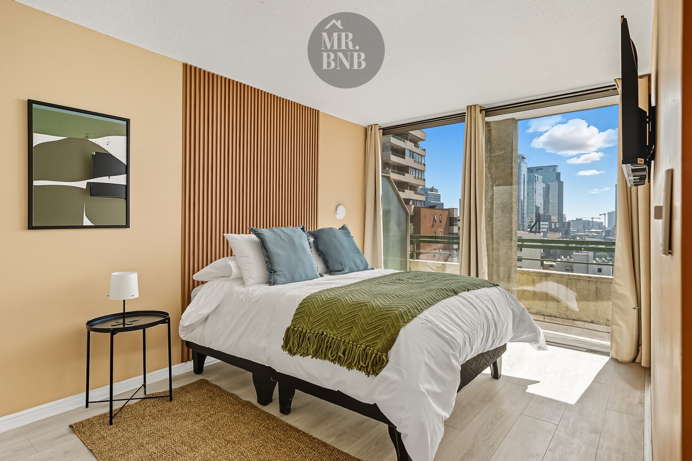 Dormitorio con cama de dos plazas, manta verde y ventanal con vista a la ciudad