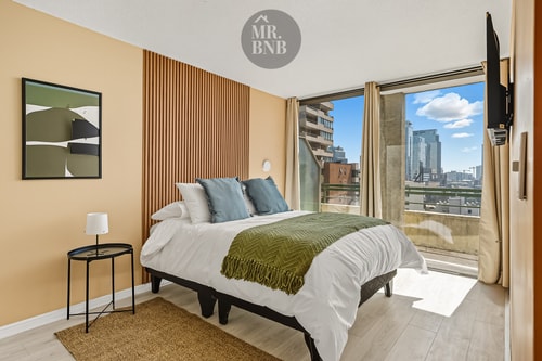 Dormitorio con cama de dos plazas, manta verde y ventanal con vista a la ciudad