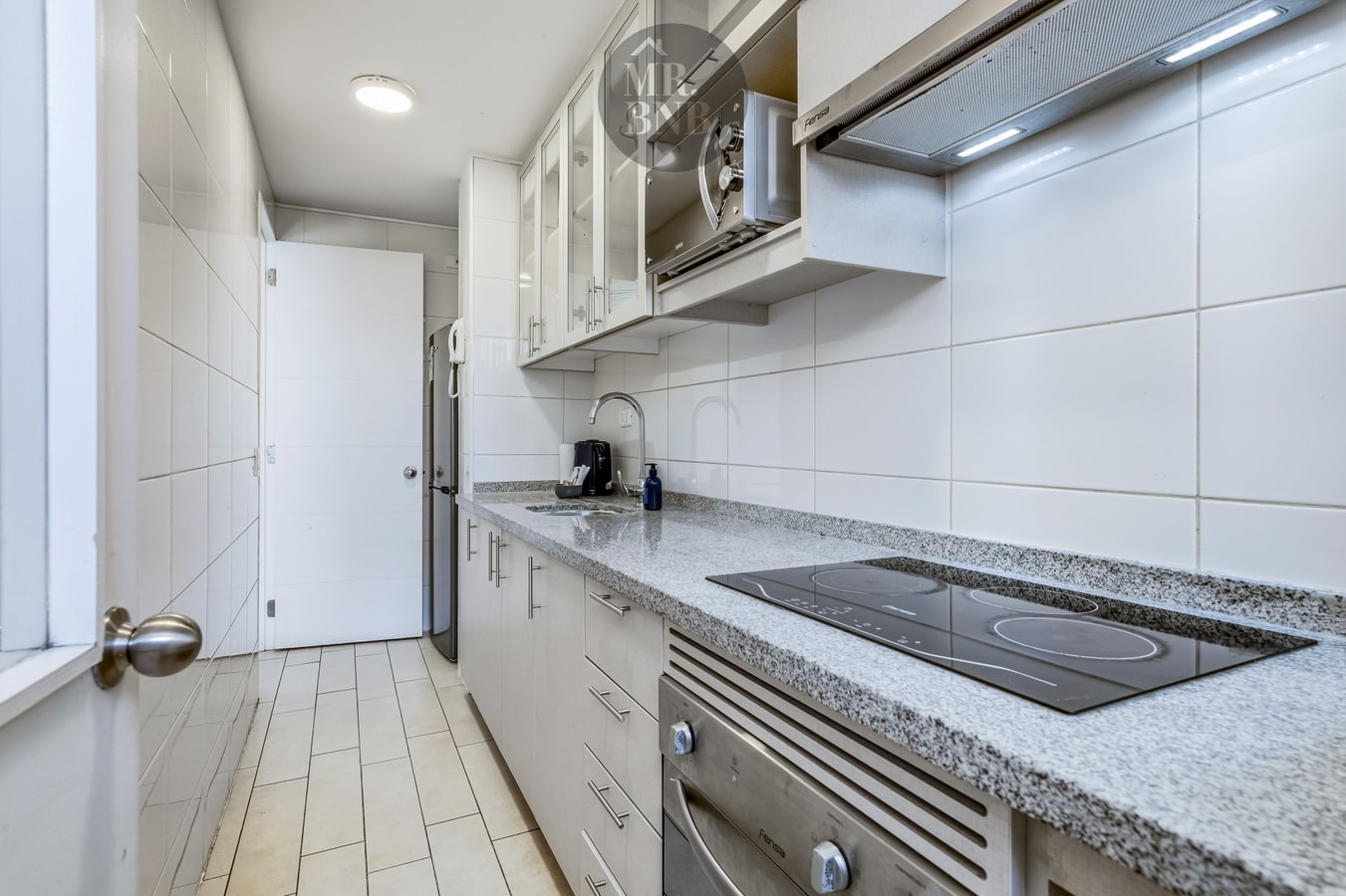 Cocina con encimera eléctrica, horno y muebles blancos.