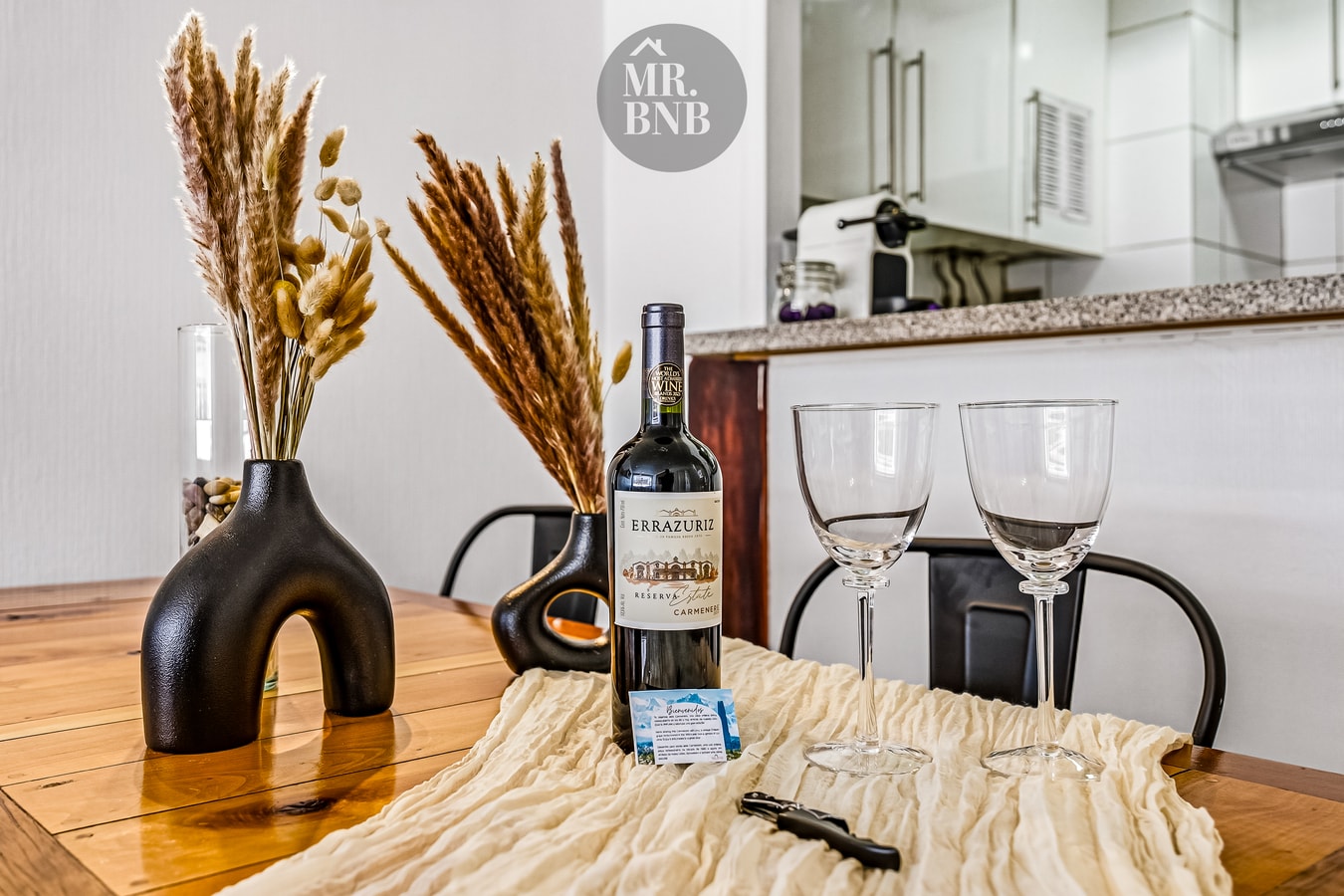 Detalle de mesa con botella de vino, copas y flores secas, con la cocina al fondo