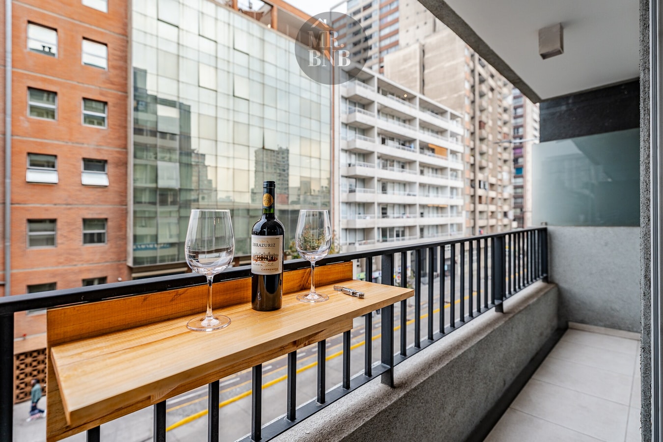 Balcón propio con mesa alta, botella de vino y copas, vista hacia la avenida y edificios.